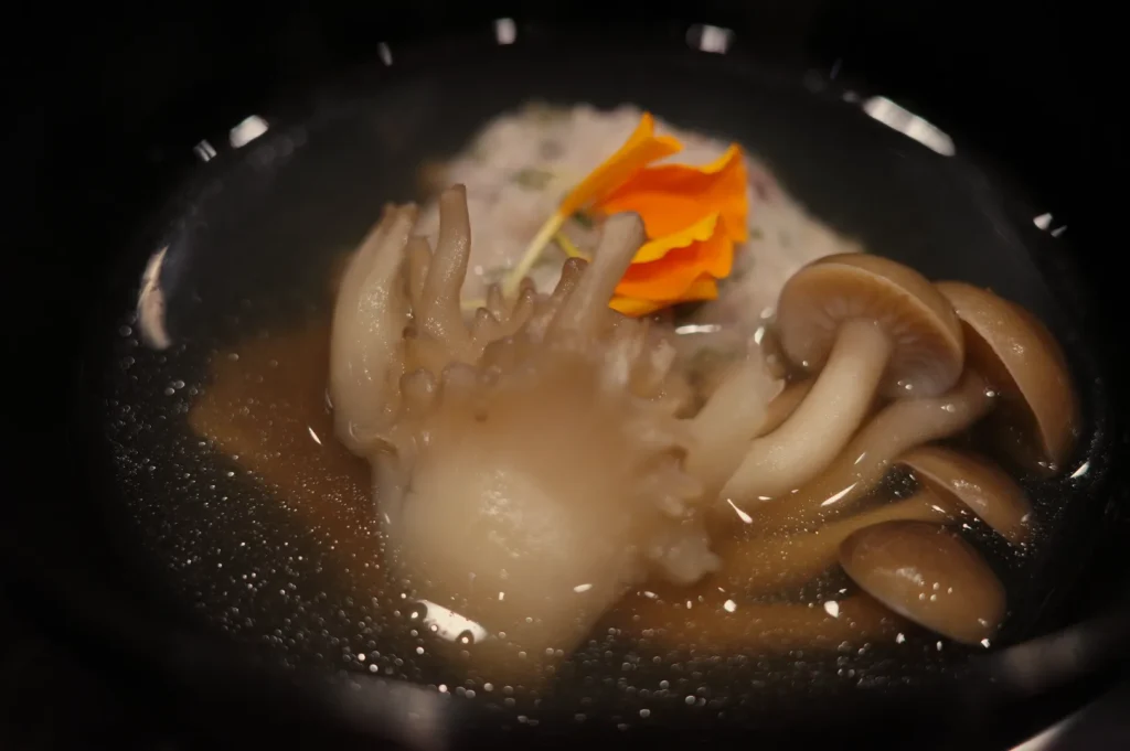 A hand-shaped piece of dough simmers in a dark pot with brown mushrooms and a bright orange garnish, creating an eerie, surreal mood.