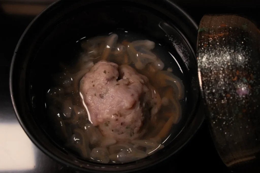Overhead view of a dark pot with pale pink, herb-speckled meat in brown broth and sliced onions; a glass lid rests beside it, creating a subdued kitchen scene.