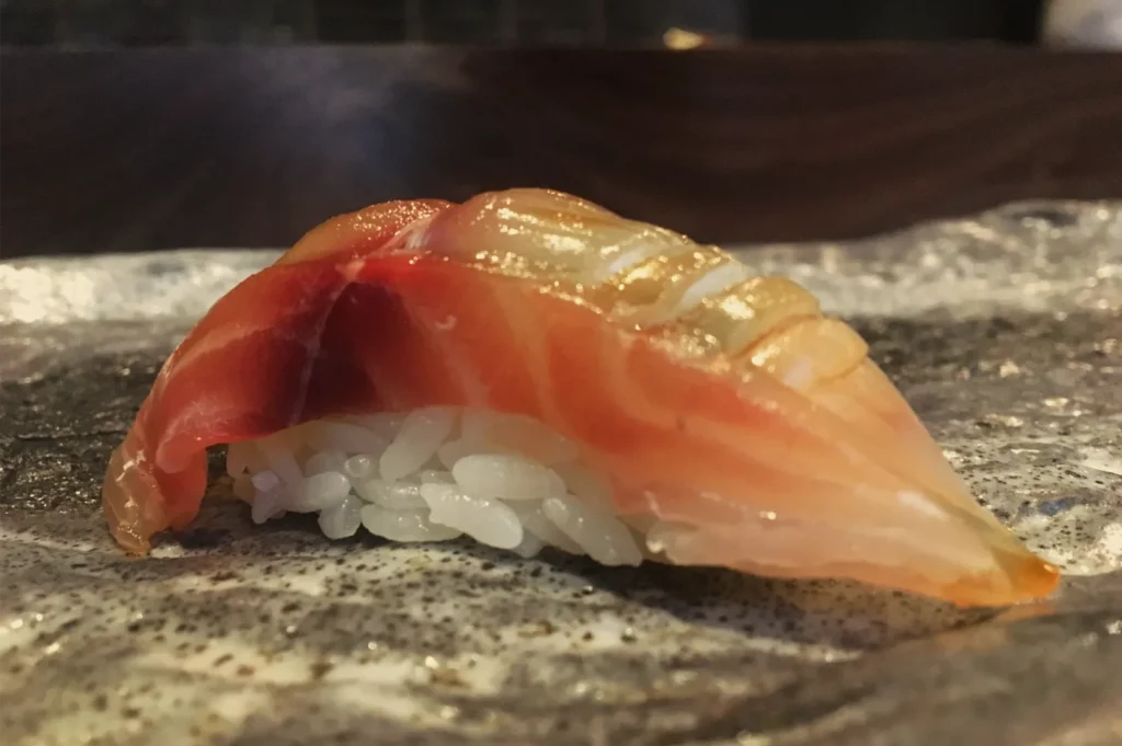 Close-up of glossy orange salmon nigiri sushi atop compact white rice on a stone plate, with a warm, appetizing restaurant mood.