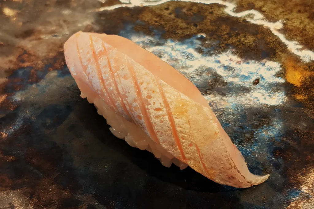 A single piece of salmon nigiri sushi rests on a dark, wet stone surface, its pale orange fish glistening in soft light for an elegant, appetizing mood.