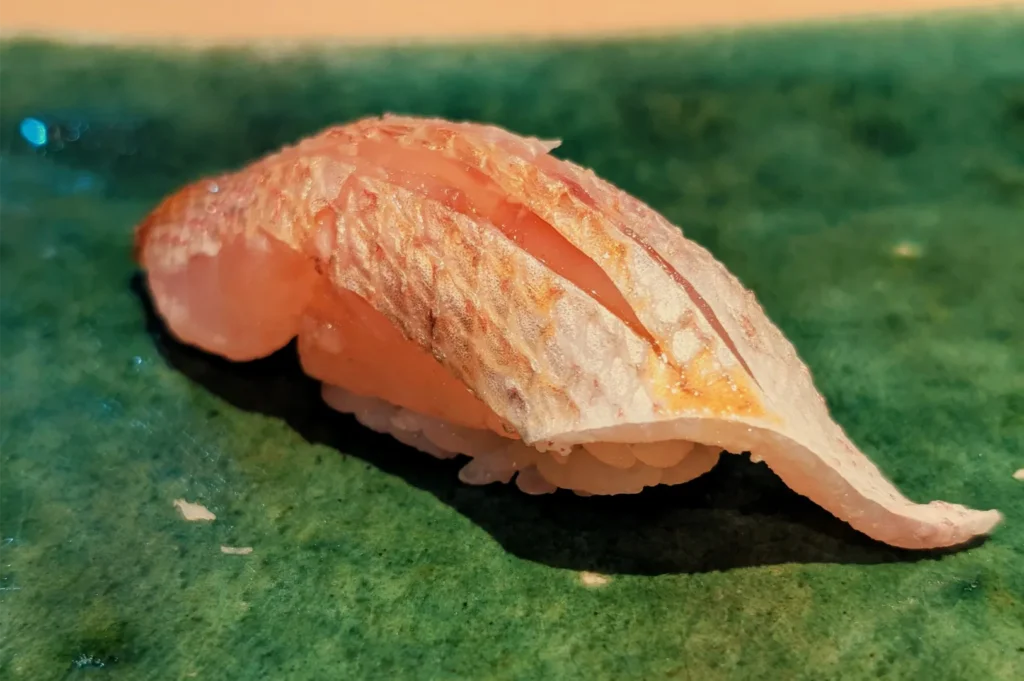 Close-up of salmon nigiri sushi, with a glossy orange fish slice draped over rice on a dark green plate; warm lighting gives the food an inviting look.