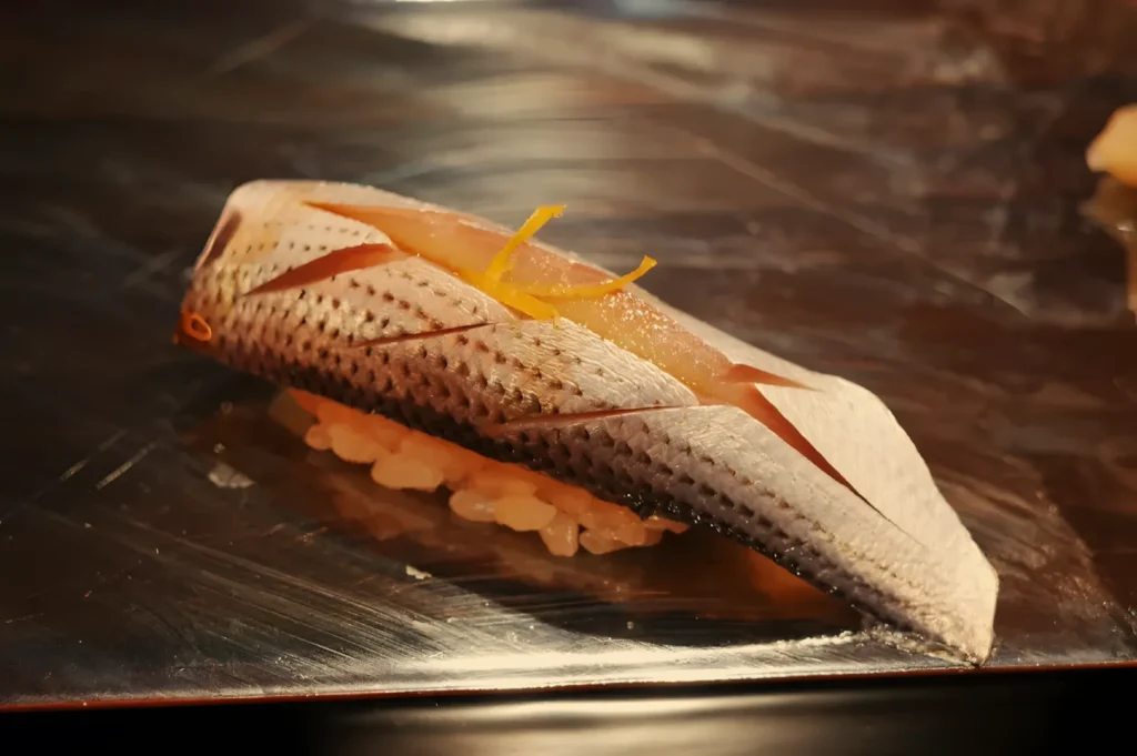 An artfully plated piece of nigiri sushi with scored fish draped over rice and yellow garnish, resting on a dark reflective platter in warm light.