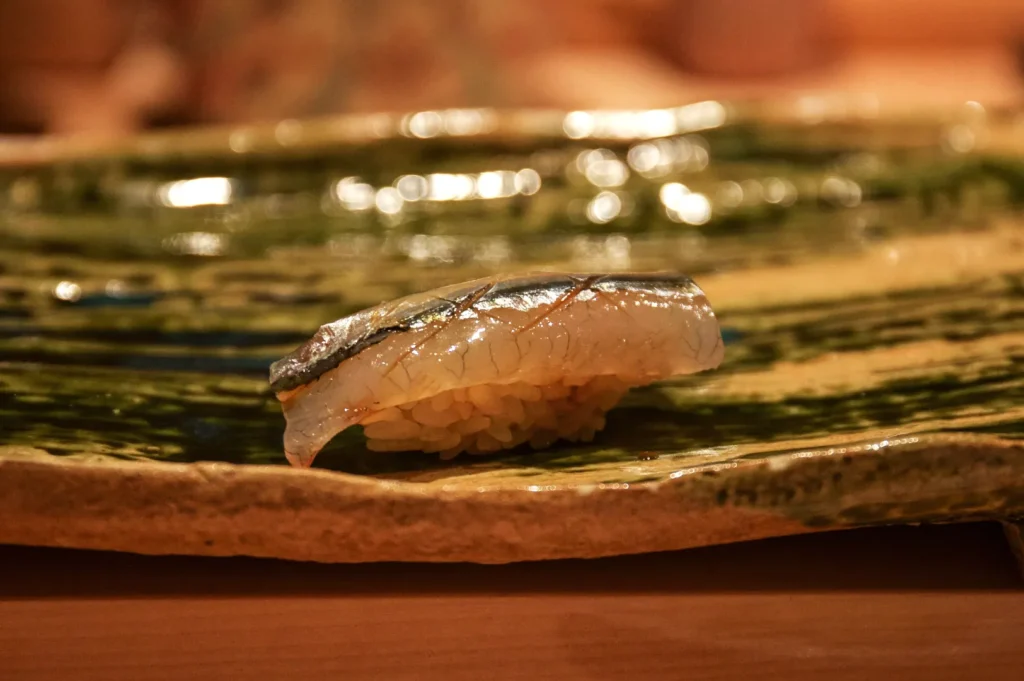 A piece of salmon nigiri sushi rests on a rustic ceramic plate, with glossy fish over compact rice; warm, dim lighting gives the close-up an intimate feel.