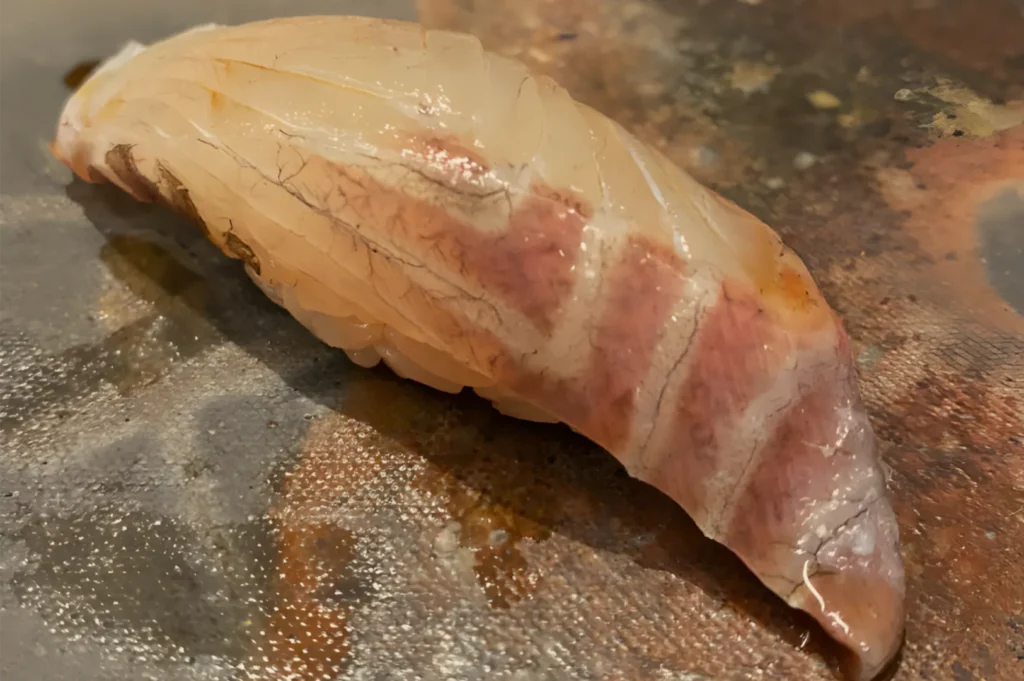 Close-up of a pale, glossy piece of raw seafood with pink marbling resting on a textured cutting board; the scene feels quiet and culinary.