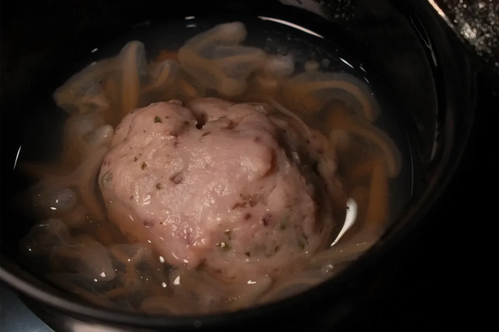 A large pale pink, herb-speckled meatball sits in brown broth with sliced onions inside a dark slow cooker, suggesting a hearty home-cooked meal.