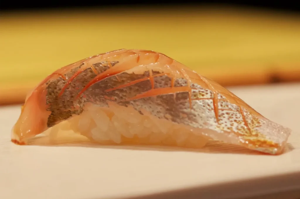 Close-up of a single piece of nigiri sushi, topped with translucent orange fish and delicate scored lines, resting on a pale plate against a warm yellow background.