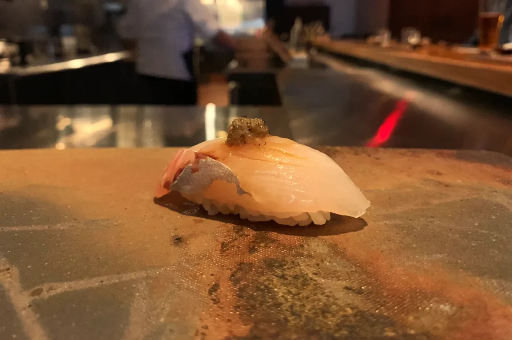 White-fish nigiri sushi with a small garnish rests on a stone counter in a dim, warmly lit restaurant, creating an intimate dining mood.