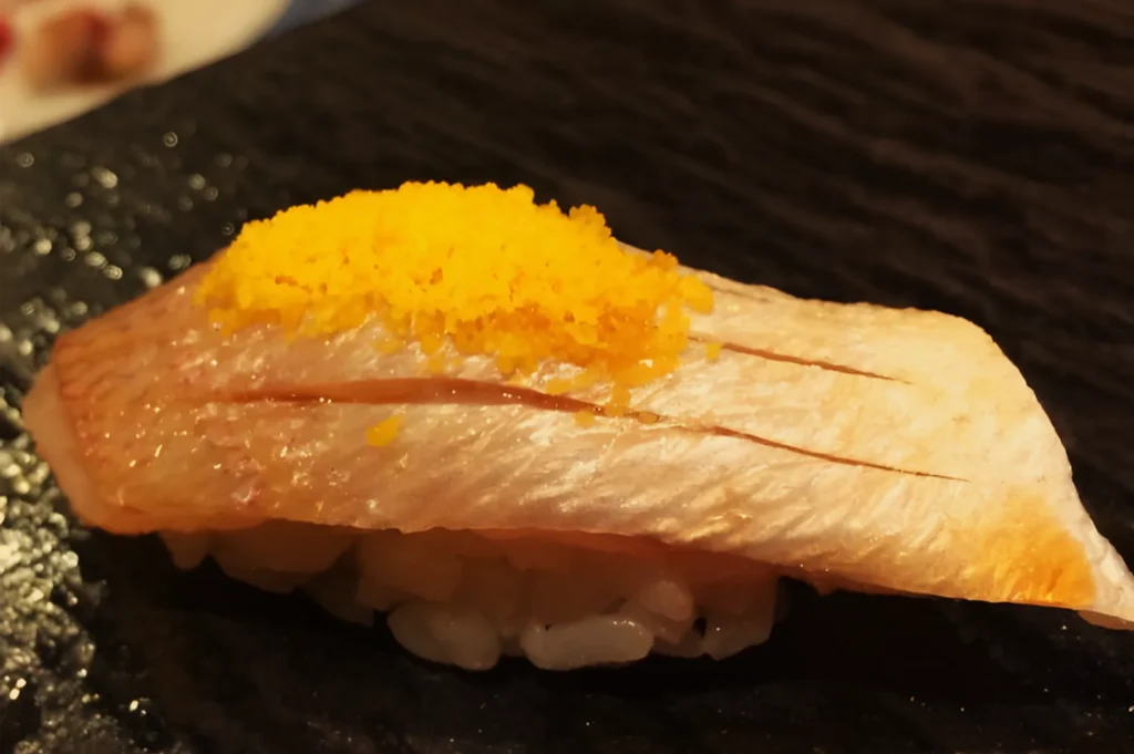 Pale fish nigiri topped with bright orange grated garnish sits on a textured black plate, its warm lighting creating an elegant, appetizing mood.
