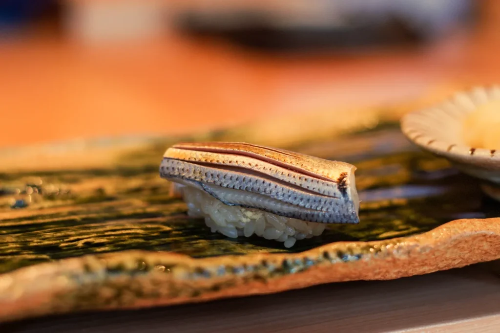 A single piece of nigiri sushi rests on a dark green leaf beside a plate, warmly lit against a softly blurred orange background, creating an inviting mood.