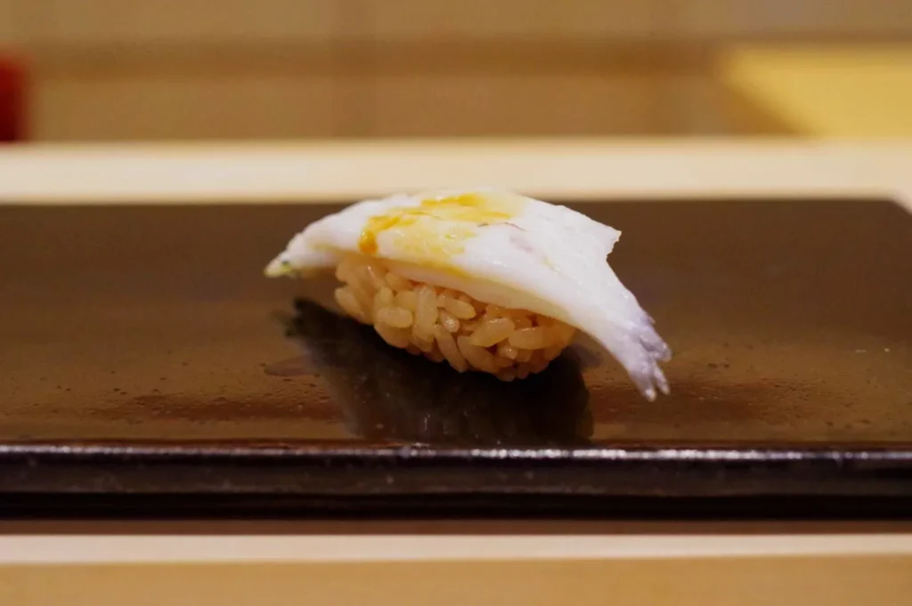 A single piece of delicate white fish nigiri rests on a dark wooden plate, topped with a light yellow garnish in a calm, minimalist sushi setting.