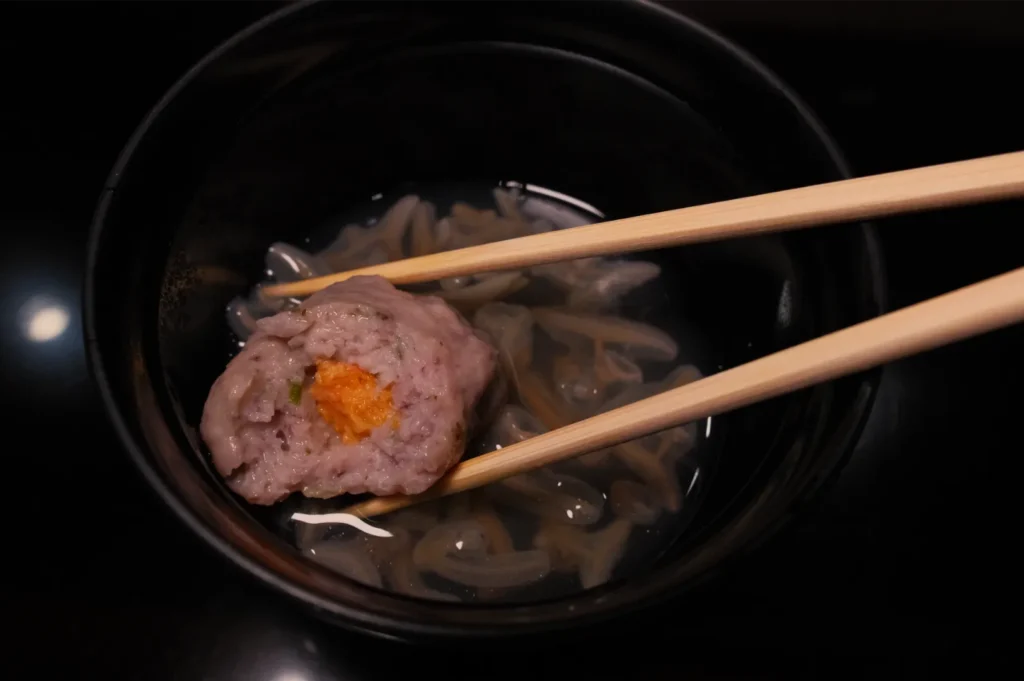 Overhead view of a black bowl of clear broth with noodles and a pink meatball topped with orange garnish, held by wooden chopsticks; a warm, appetizing meal.