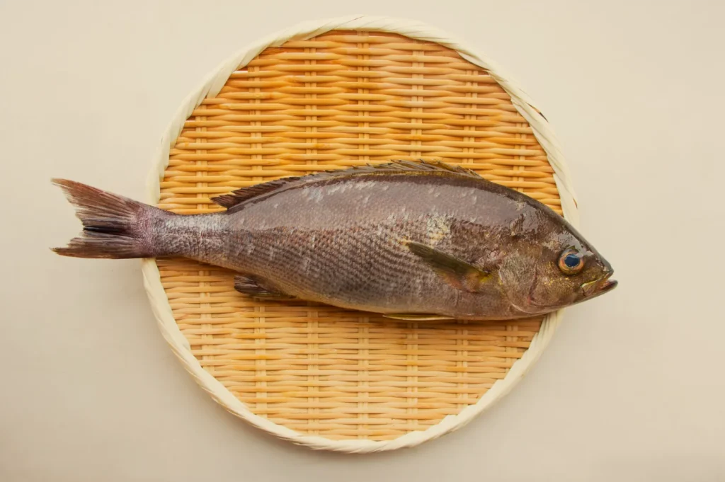A whole raw fish lies horizontally on a round woven bamboo tray against a plain beige background, with its tail left and head right; the mood is quiet and rustic.