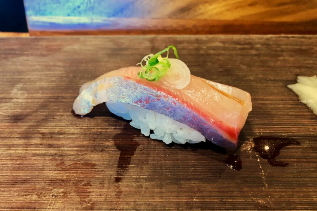 A colorful piece of nigiri sushi with pale pink fish and a green garnish rests on a dark wooden counter, creating an appetizing restaurant scene.