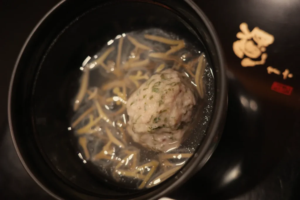 Overhead view of a black pot with clear broth, thin noodles, and a large herb-topped meatball; warm lighting gives the dish a cozy feel.