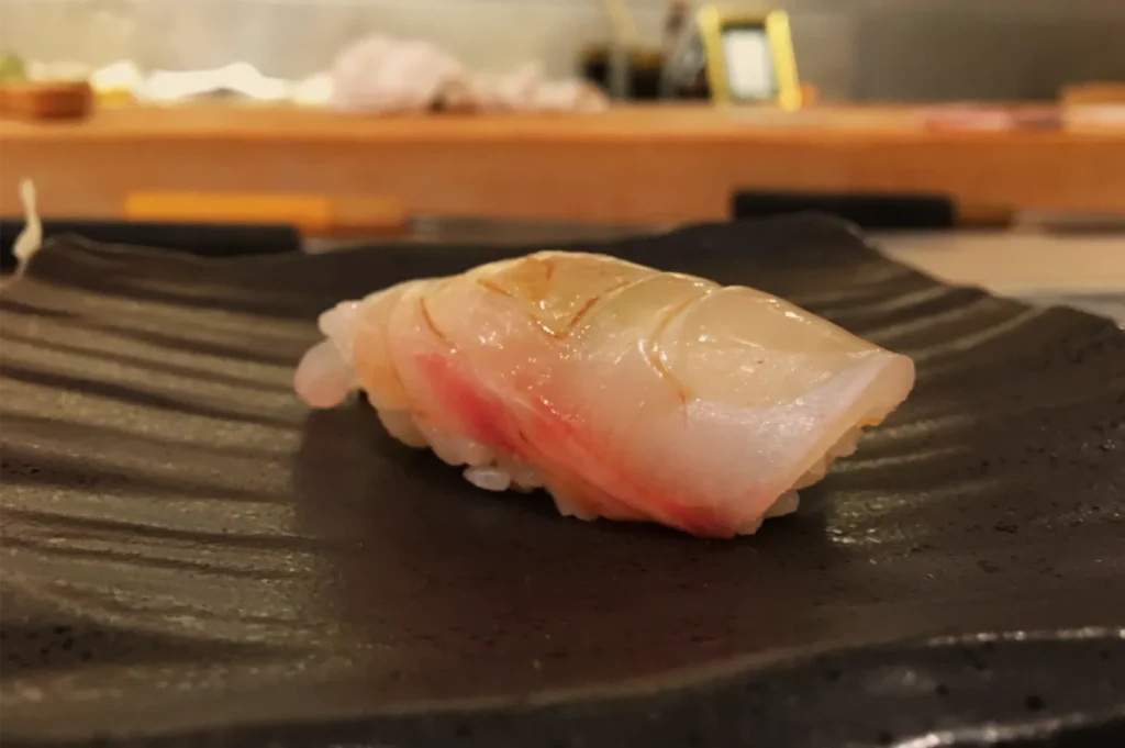 A glossy slice of pale pink and white raw fish nigiri sushi rests on a dark ridged plate at a sushi counter, creating an elegant, appetizing mood.