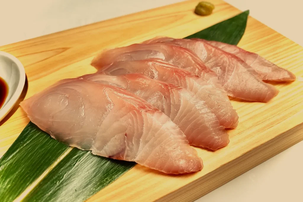 Fresh translucent slices of pale pink sashimi are arranged on a green leaf atop a wooden board, with soy sauce and wasabi nearby; the presentation feels elegant and appetizing.