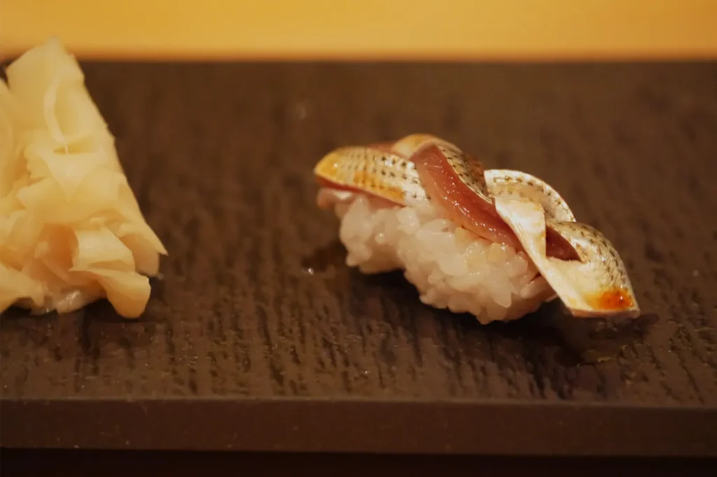 Close-up of nigiri sushi topped with glossy fish and a thin strip of garnish on a dark wooden plate, with pickled ginger beside it; warm, intimate dining mood.