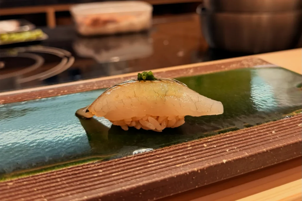 A single piece of translucent white fish nigiri with a green herb garnish rests on a glossy ceramic plate at a sushi counter, creating a refined, calm mood.