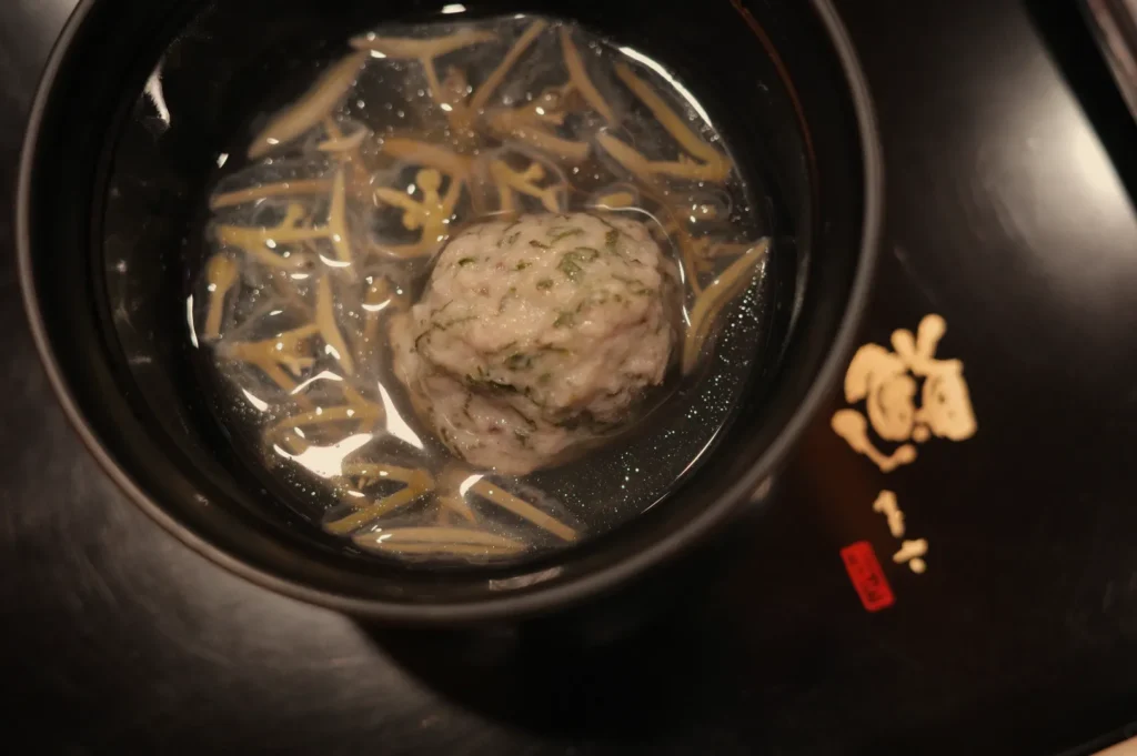 A warm bowl of clear broth with a herb-speckled meatball and thin noodles sits on a dark table, accompanied by small garnishes and a red label.