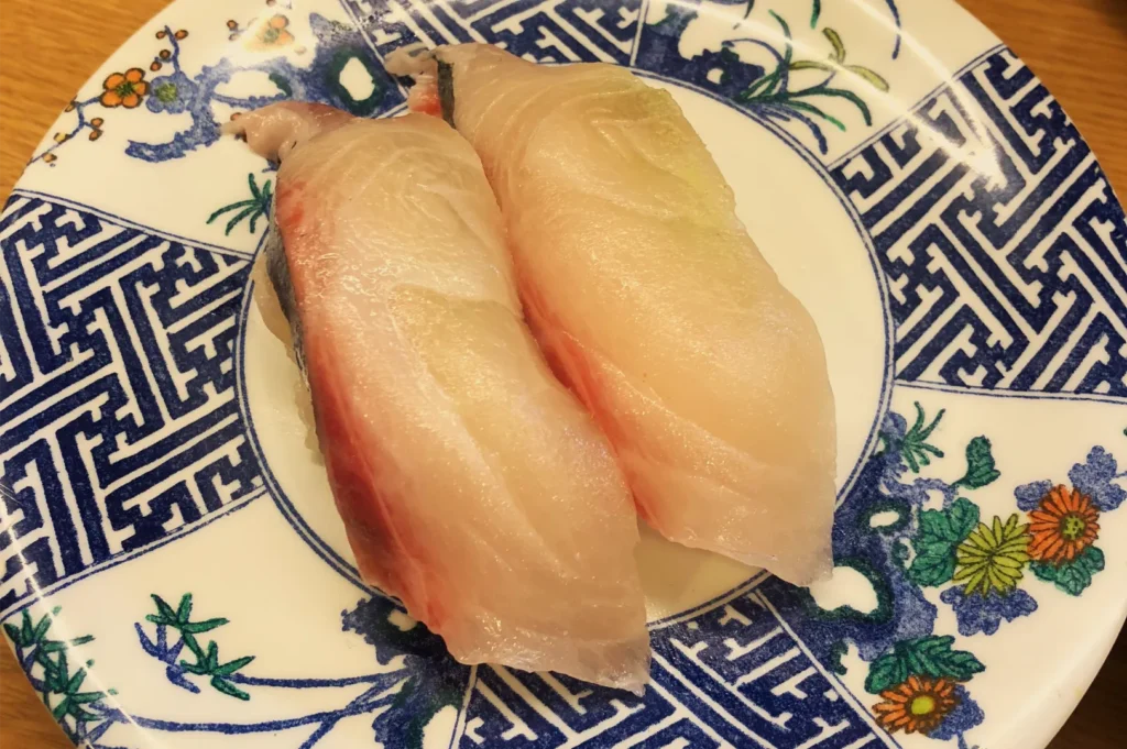 Two pieces of pale, translucent white-fish nigiri sushi sit on an ornate blue-and-white floral plate, creating an appetizing close-up.