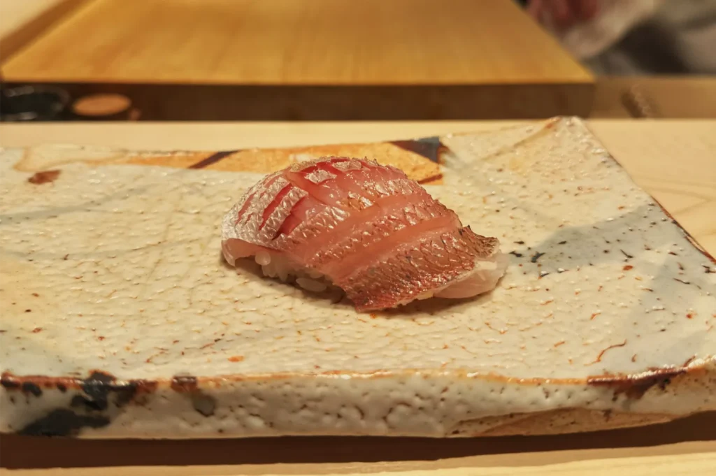 A piece of glossy red fish nigiri rests on a rustic, cream-colored ceramic plate at a sushi counter, creating a calm, refined dining scene.