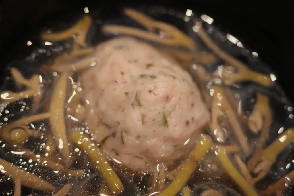 Close-up of a pale, round dumpling in a glossy brown broth, surrounded by noodles and small vegetables; the warm, rustic dish looks hearty and comforting.