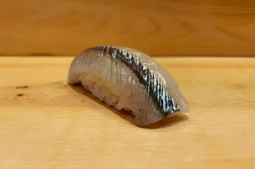 A single piece of silver-skinned fish nigiri rests on a pale wooden sushi board, its glossy surface catching the light in a quiet, minimalist setting.