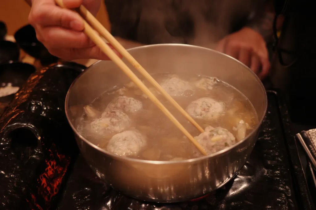 Hands use wooden chopsticks to stir meatballs in a steaming metal pot over a tabletop burner, capturing the warm, communal feel of a hot-pot meal.