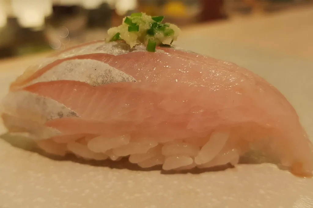 Close-up of glossy pink tuna nigiri sushi over compact white rice, topped with a small mound of grated garnish and green herbs; warm, inviting restaurant lighting.