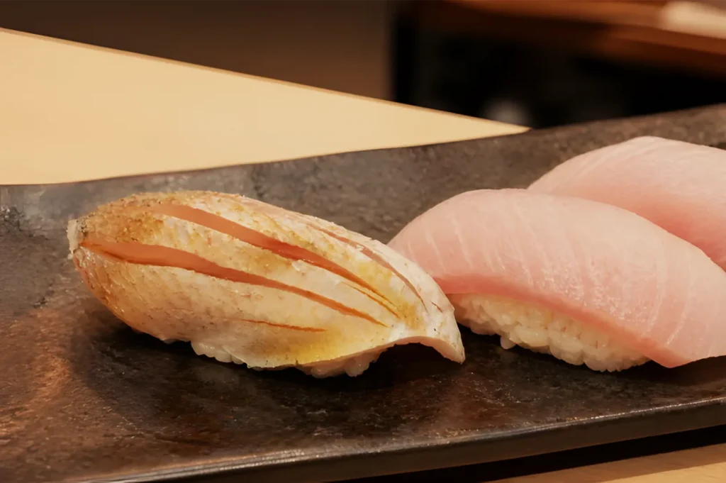 Two pieces of nigiri sushi sit on a dark stone plate: browned, scored fish on the left and pale pink tuna on the right, warmly lit and inviting.