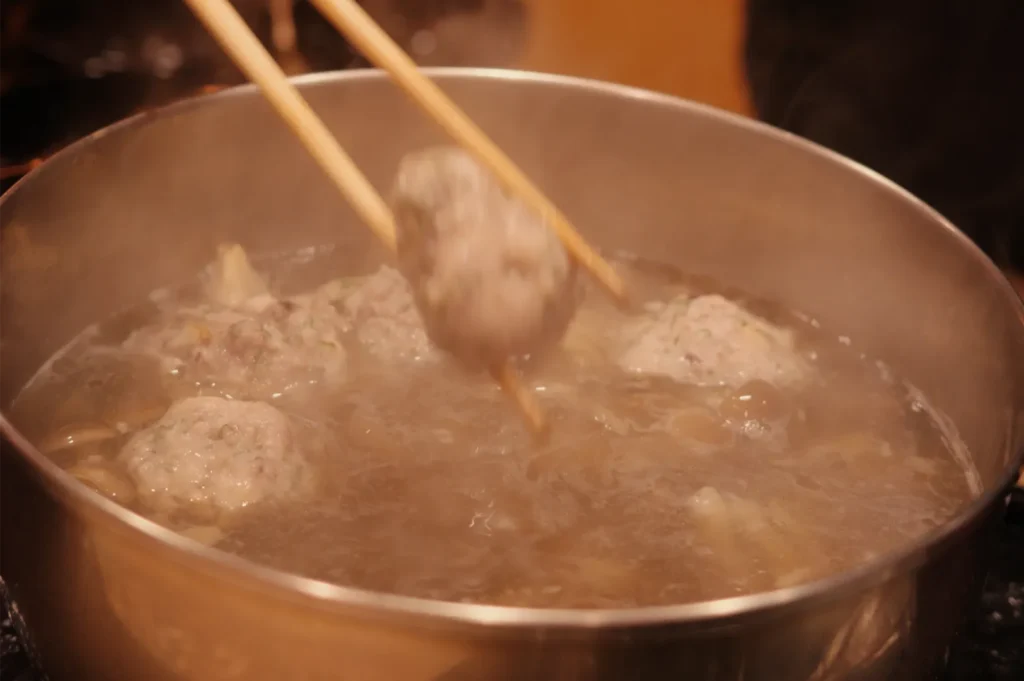 Chopsticks lift a meatball from a steaming pot of simmering broth, with more meatballs cooking inside; the warm, cozy scene suggests home cooking.